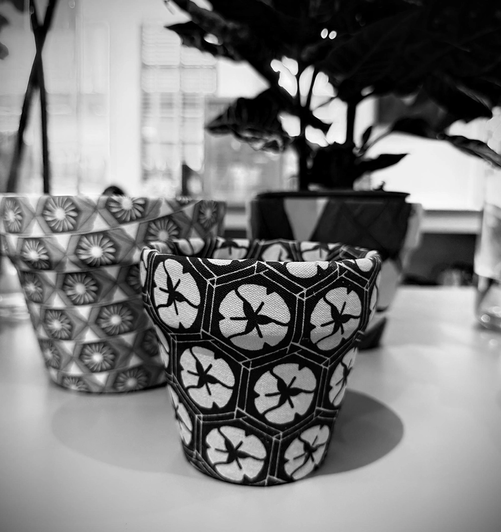 pot-new-4 Black and white photo of a pot covered in a hexagon flower print, in front of a potted plant.